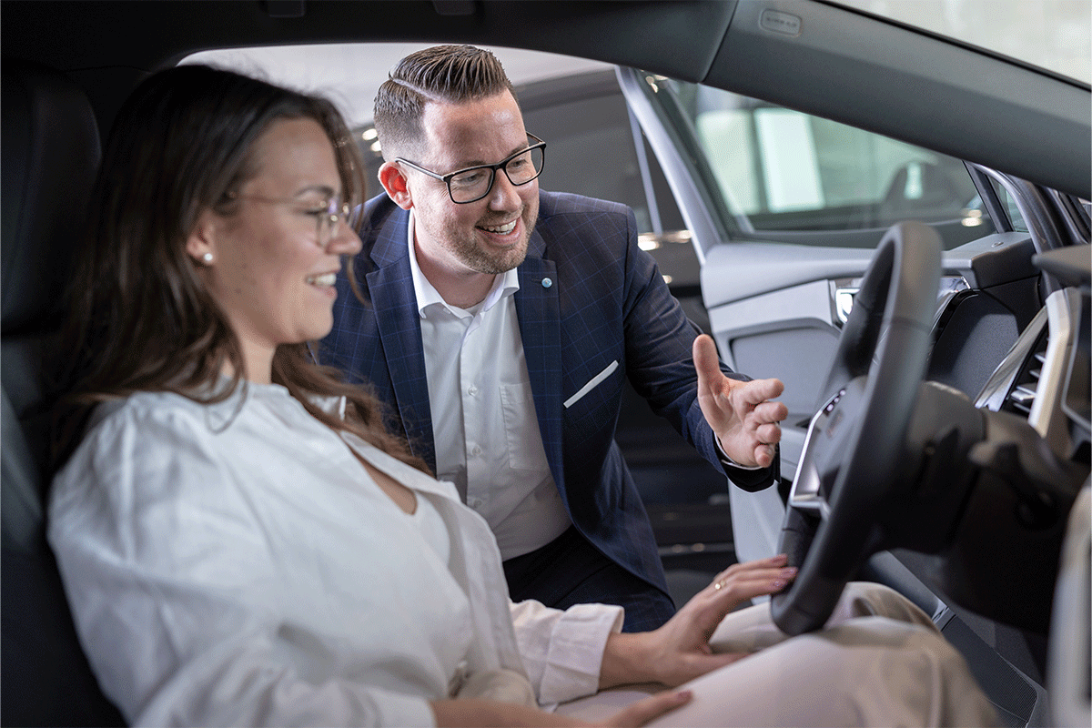 Verkäufer erklär in Fahrzeug sitzender Frau die Funktionen des Autos