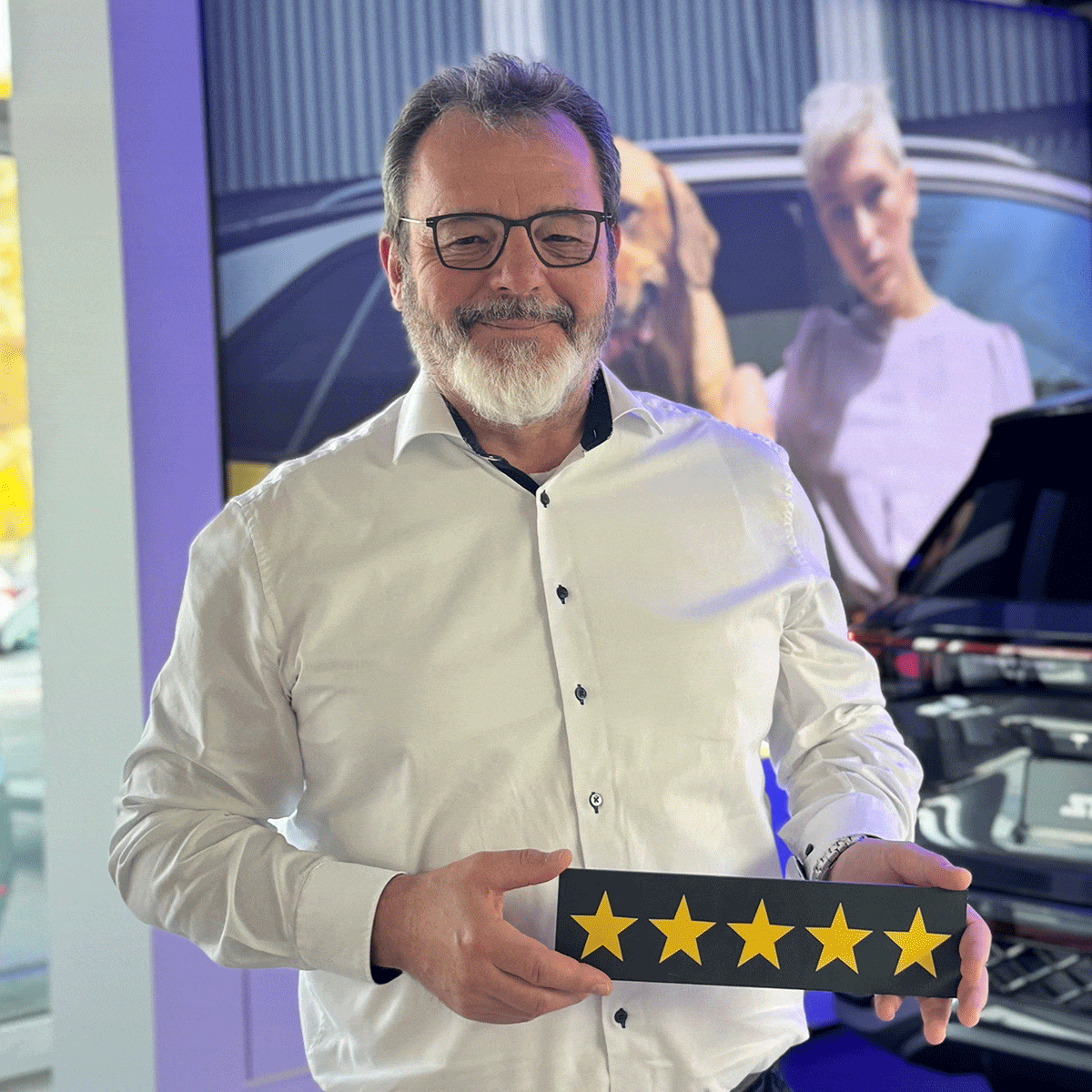 Neuwagen Verkäufer Frank Meyer lächelt in die Kamera und hält ein Schild mit fünf Sternen in der Hand.