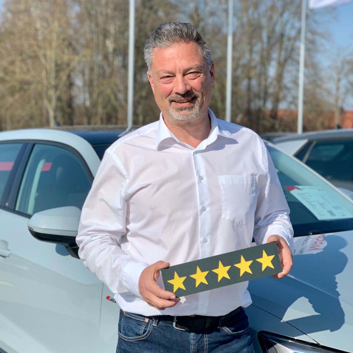 Gebrauchtwagen Verkäufer Thomas Lechner lächelt in die Kamera und hält ein Schild mit fünf Sternen in der Hand