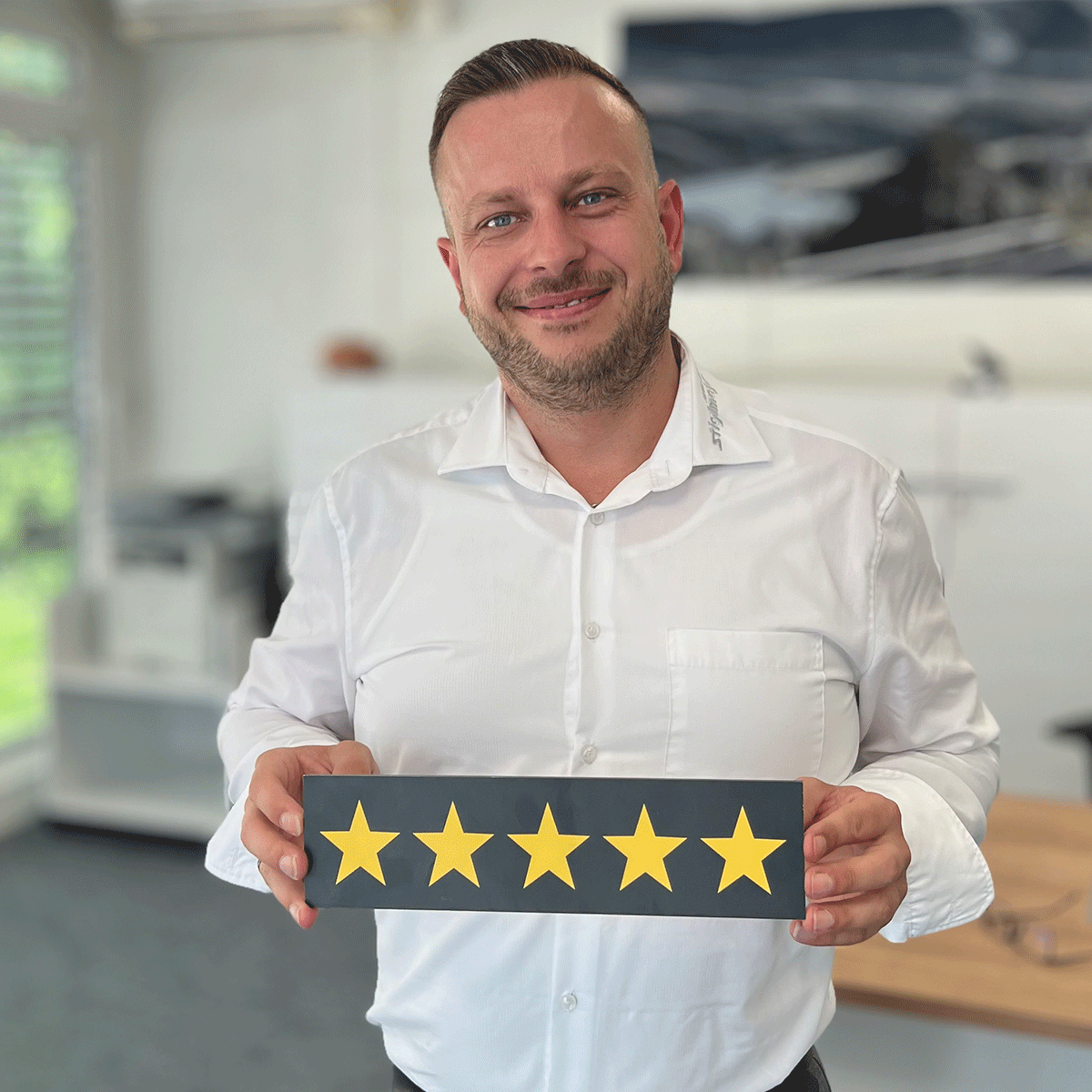 Gebrauchtwagen Verkäufer Marco Zierer lächelt in die Kamera und hält ein Schild mit fünf Sternen in der Hand.
