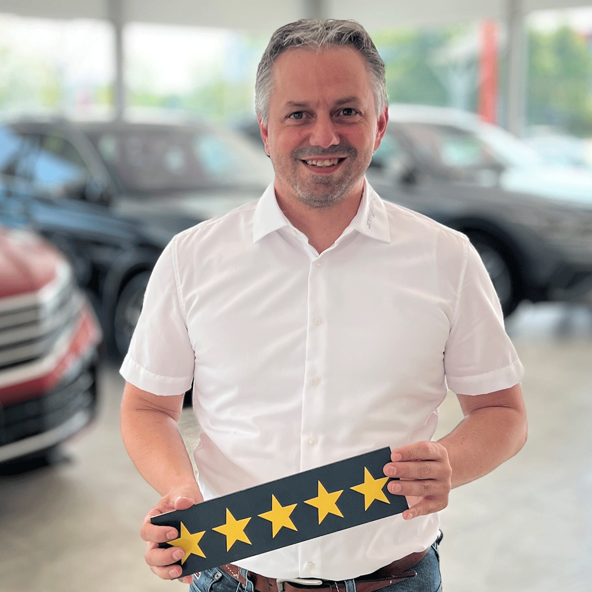 Gebrauchtwagen Verkäufer Michael Finke lächelt in die Kamera und hält ein Schild mit fünf Sternen in der Hand.