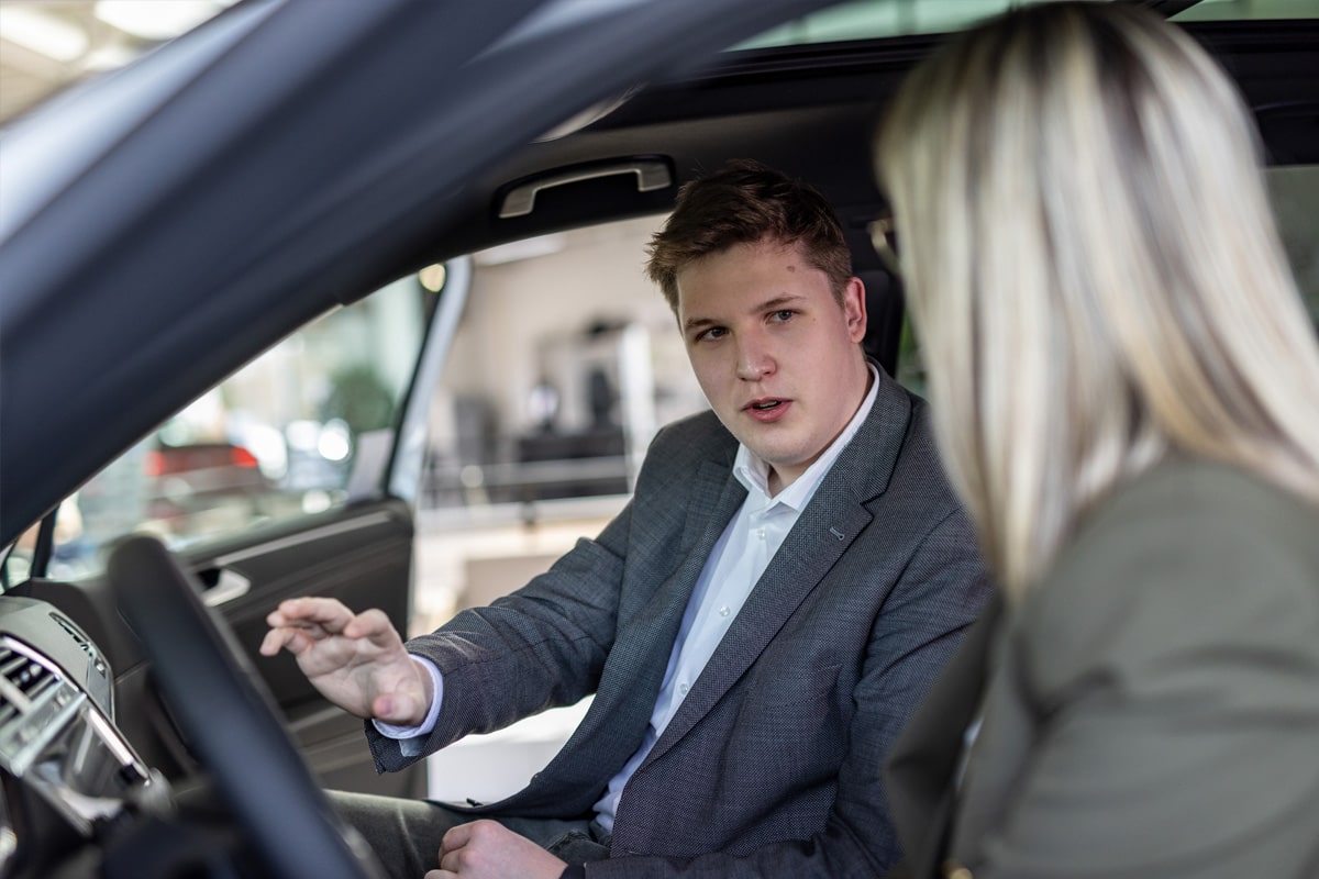 Ein Autoverkäufer sitzt mit einer Kundin in einem Neuwagen und erklärt Ihr die Fahrzeugdetails.
