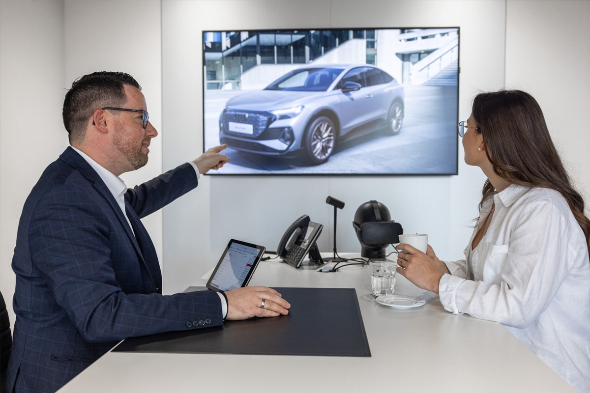 Autoverkäufer sitzt mit Kundin an einem Tisch und zeigt ihr auf einem großen Bildschirm einen Audi Neuwagen. 